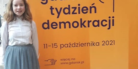 Powiększ grafikę: Kinga Zigert stoi na tle plakatu tygodnia demokracji.