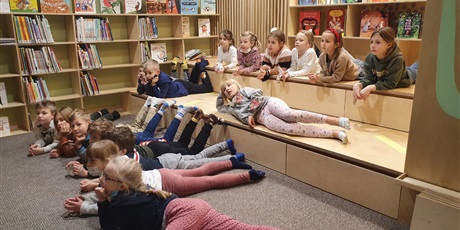 Powiększ grafikę: Na zdjęciu widać dzieci z klasy 1b, które leżą na dywanie w bibliotece i słuchają czytanej im książki.